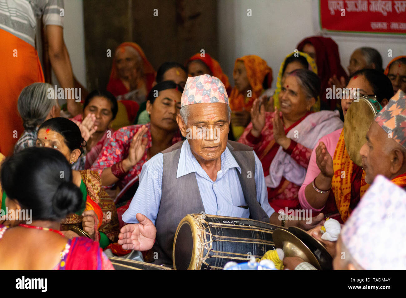 Nepalese middle aged man with traditional Dhaka topi hat playing madal drum at dance celebration surrounded by women. Stock Photo