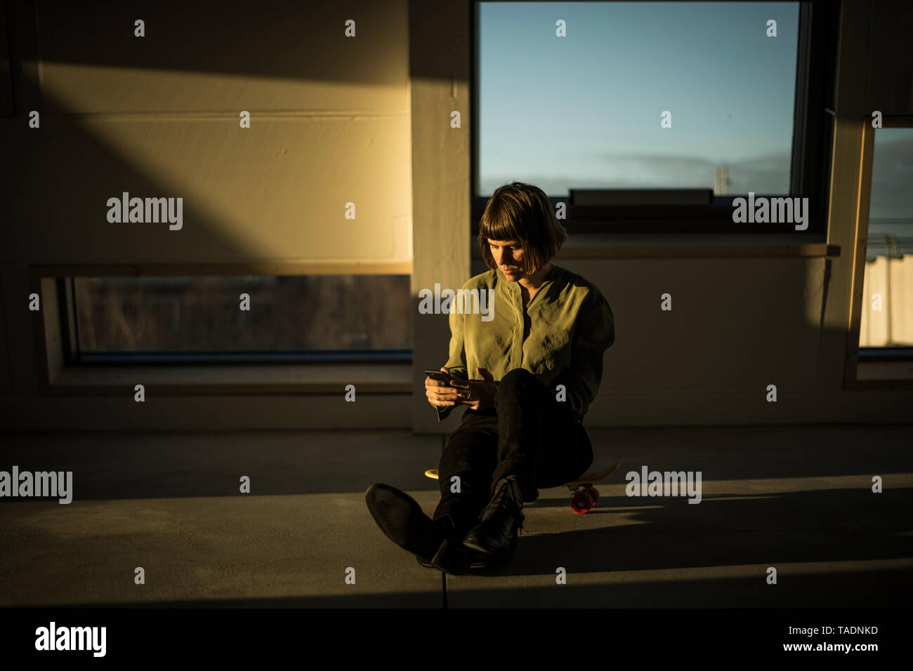 Businesswoman sitting on office floor at sunset, using smartphone Stock Photo