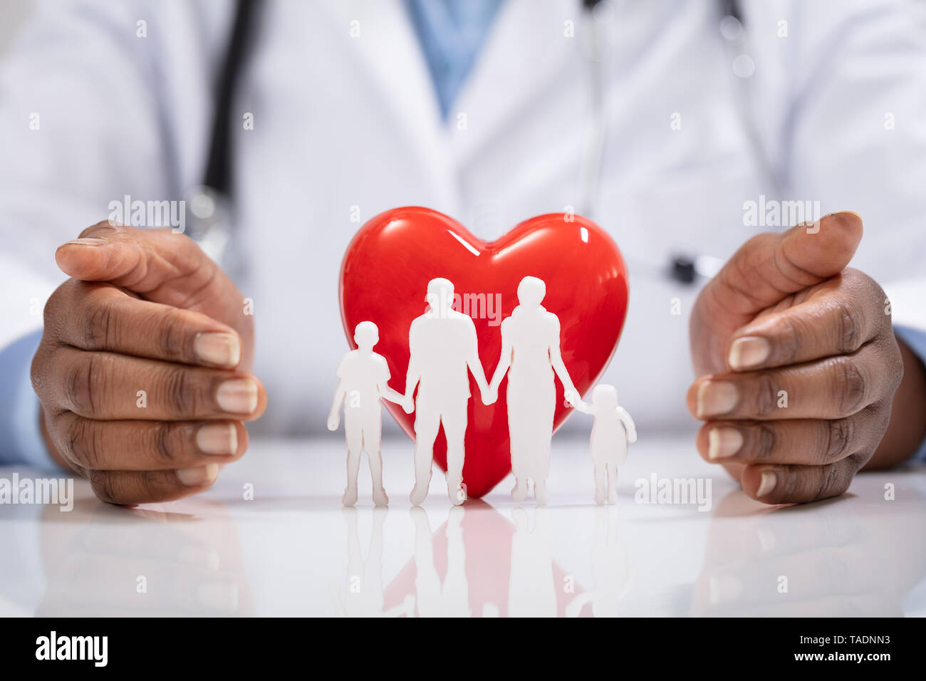 Close-up Of A Doctor's Hand Protecting White Family Paper Cut Out With Red Heart Shape Stock Photo