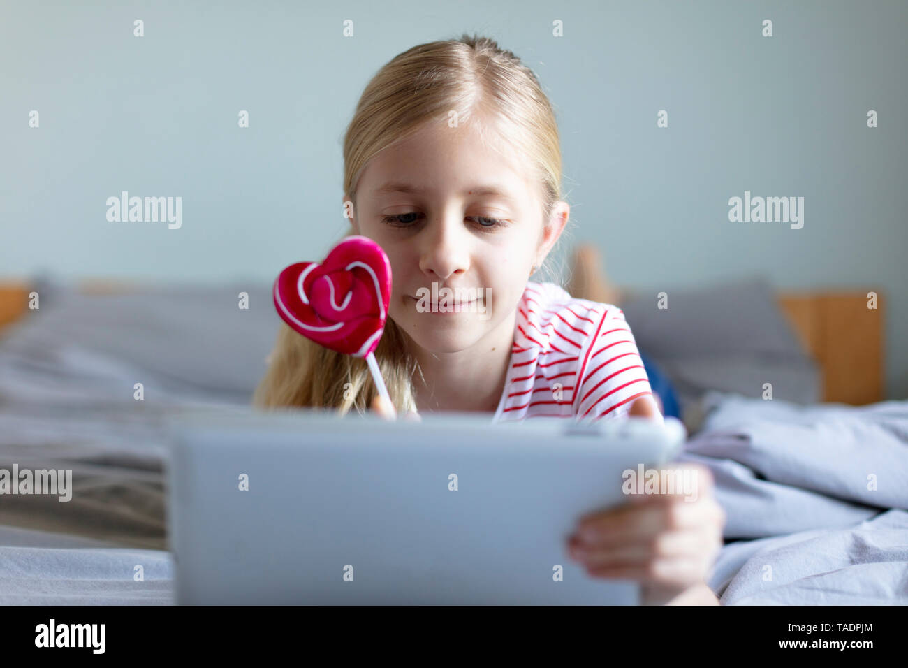 Portrait of blond girl lying on bed with lollipop using digital tablet Stock Photo