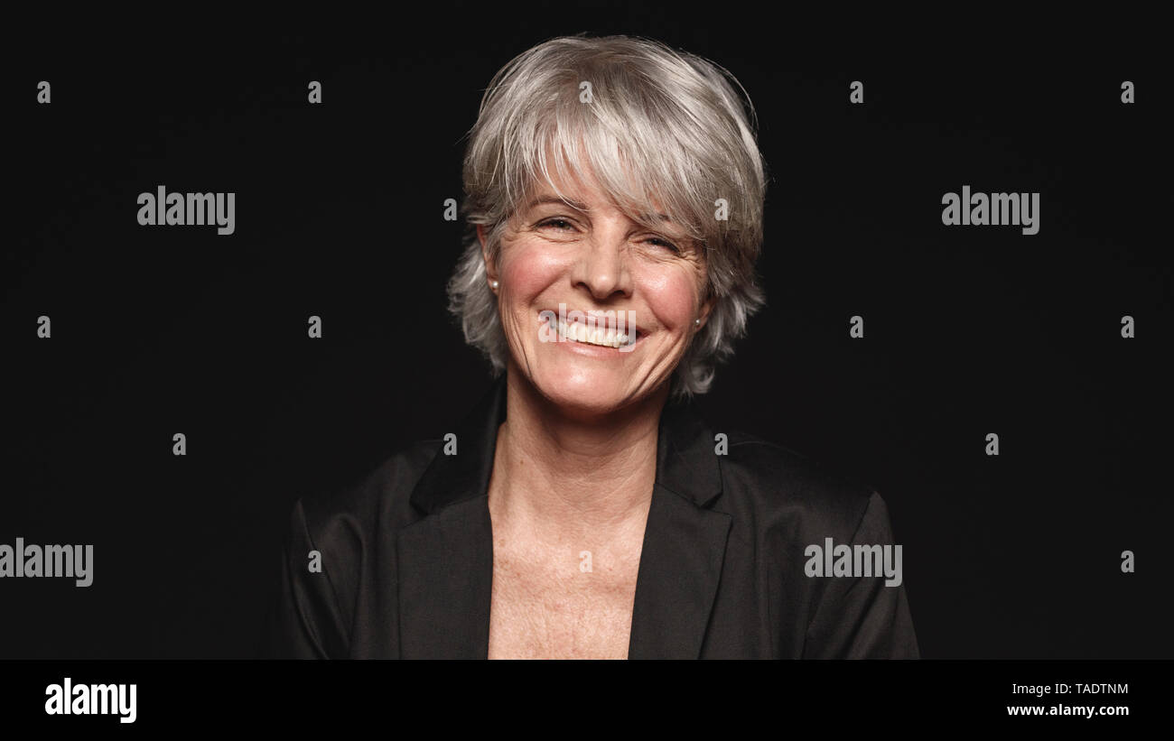 Senior woman with white hair laughing while looking at camera. Close up of smiling woman in formal wear isolated on black background. Stock Photo
