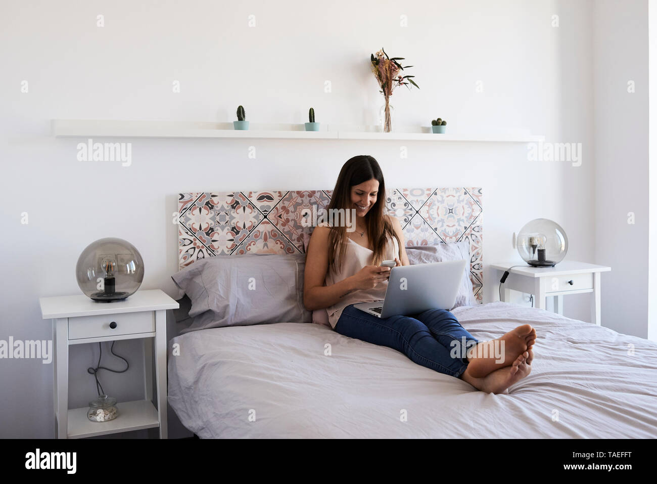 Smiling young woman sitting on bed with laptop using smartphone Stock Photo