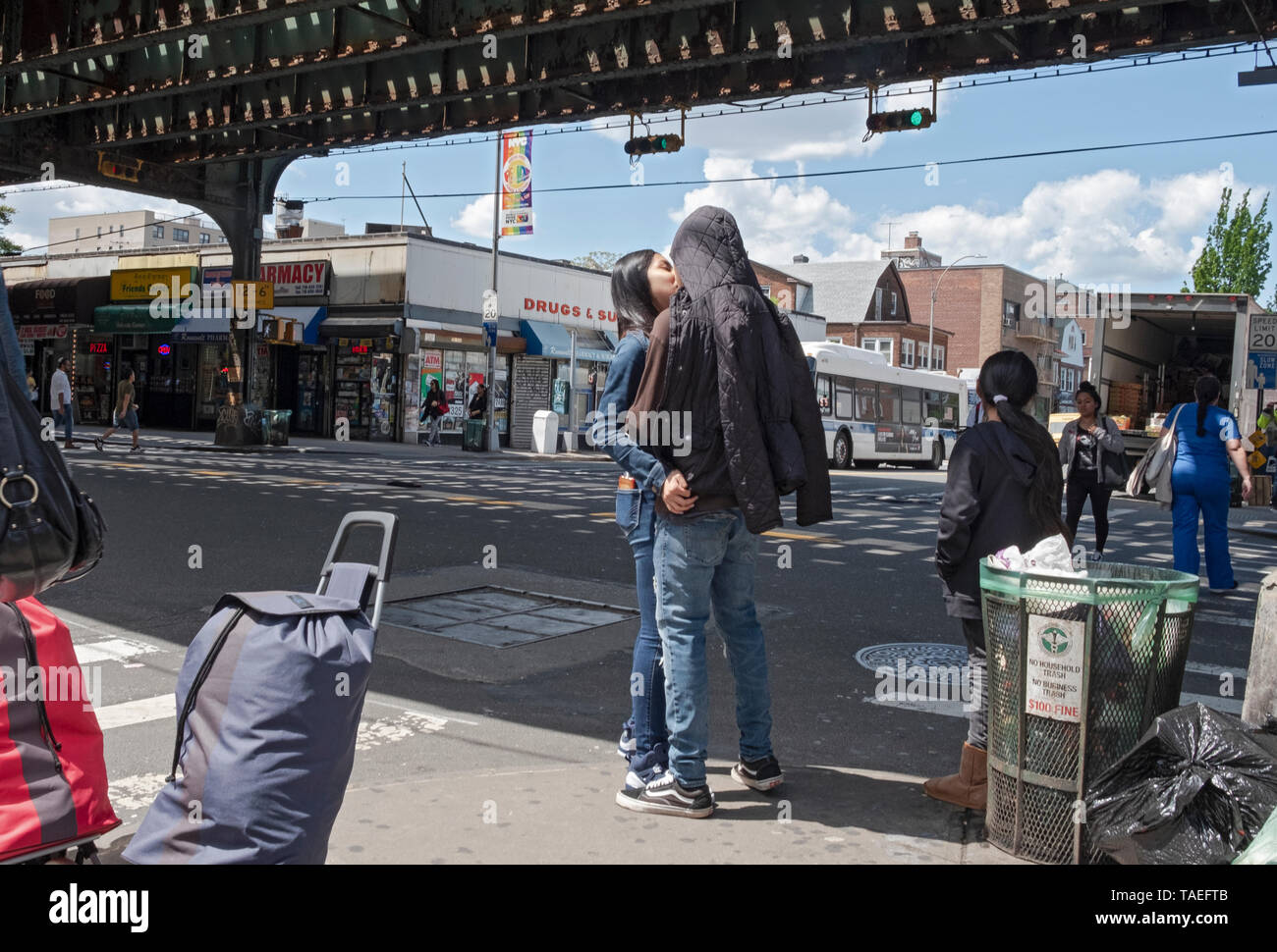 A busy street scene under the elevated subway with a couple kissing, pedestrians, luggage & trash. In Jackson Heights, Queens, New York City. Stock Photo