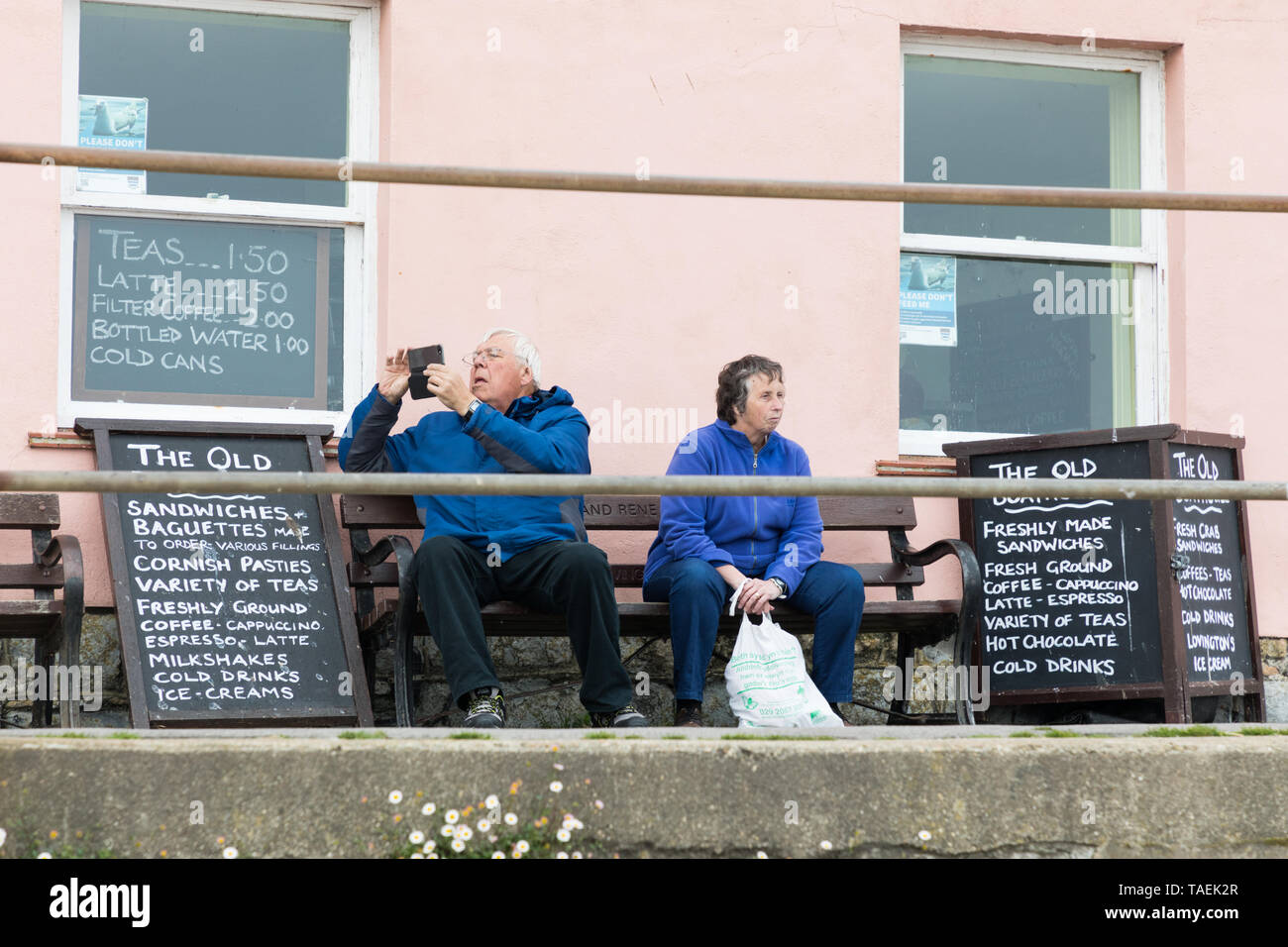 Lyme Regis, Dorset, UK: Elderly man and woman sit on a bench between blackboards listing food and drink. He looks at a mobile phone, she stares ahead. Stock Photo