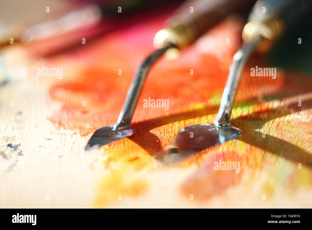 Two palette knife of different sizes lie on a colorful palette, stained with oil paints of red, yellow and orange colors. The palette knife's dirty af Stock Photo