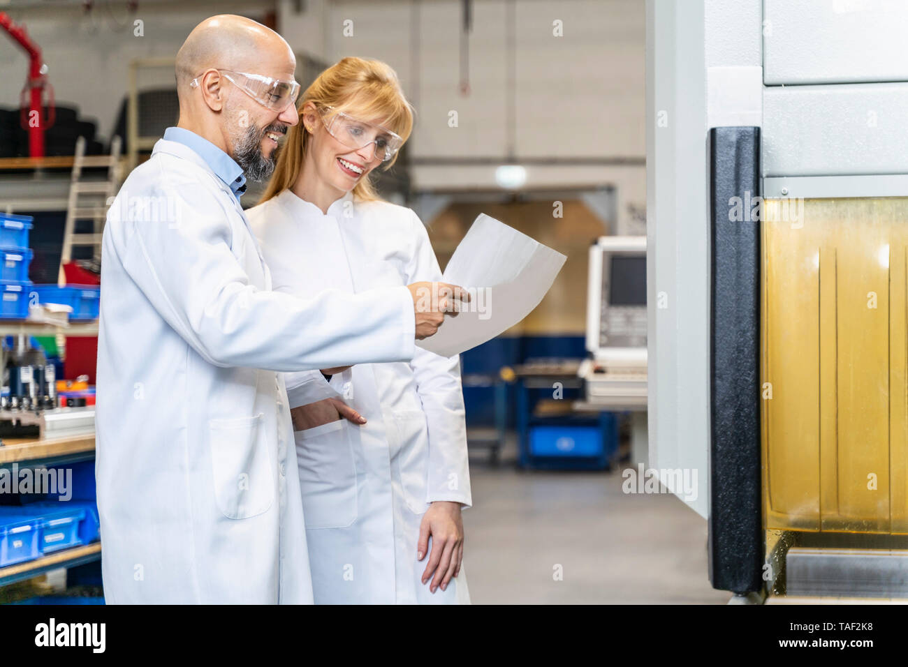 Two happy technicians wearing lab coats and safety glasses looking at plan in factory Stock Photo