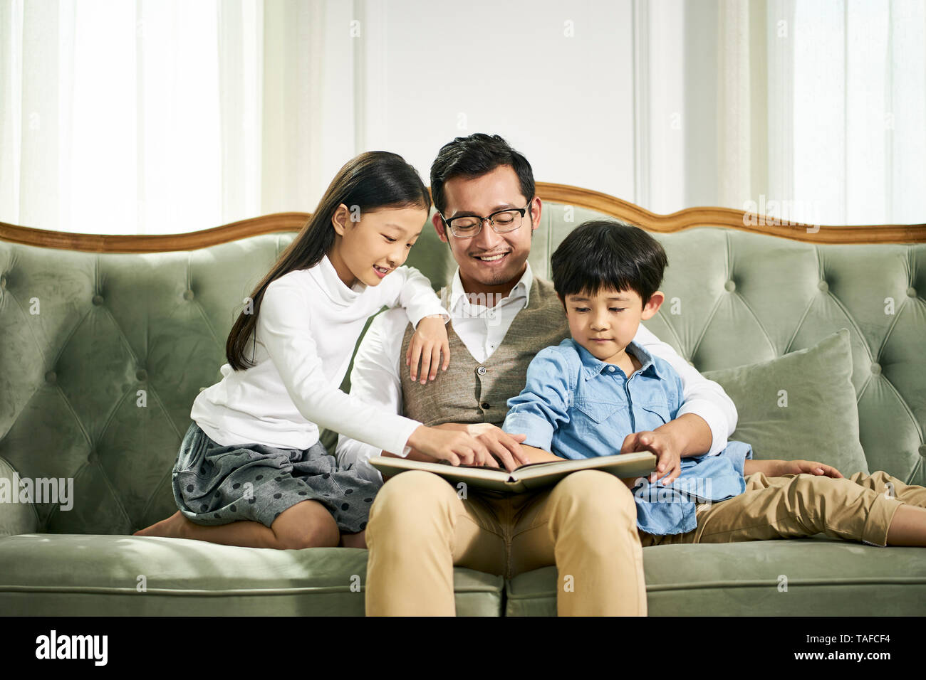 young asian father and two children sitting on couch reading book together in family living room at home Stock Photo