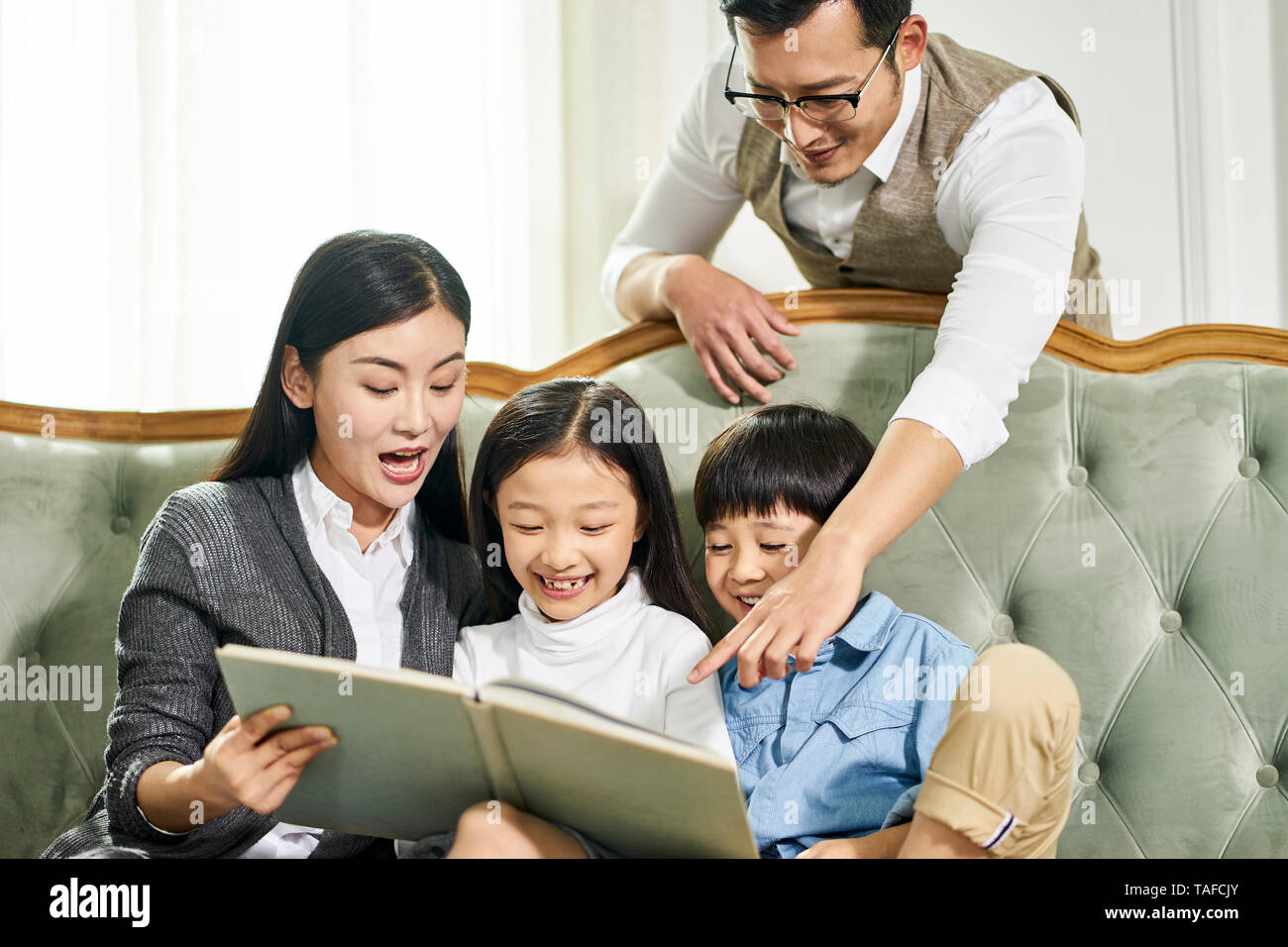 young asian parents and two children sitting on couch reading book together in family living room at home Stock Photo