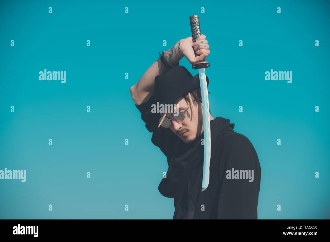 Man holding sword with metal blade on blue sky. Warrior in sunglasses, black hat and clothes. Samurai with japan katana weapon outdoors. Martial arts  Stock Photo