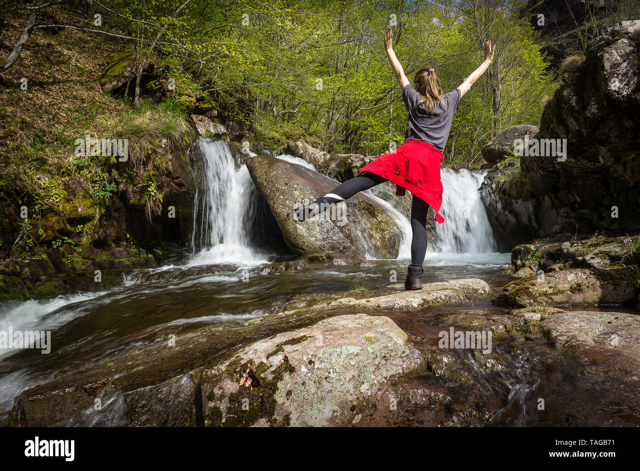 Girl with red jacket around her waist standing on one leg with hands in the air in front of scenic, waterfall on a mountain creek during early spring Stock Photo