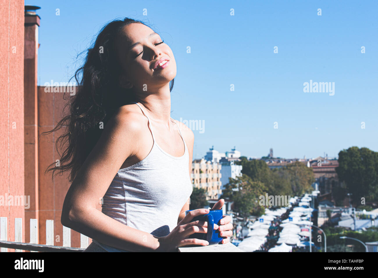 Teenager asian girl eyes closed holding mug for breakfast in day sun light, urban background view from balcony Stock Photo