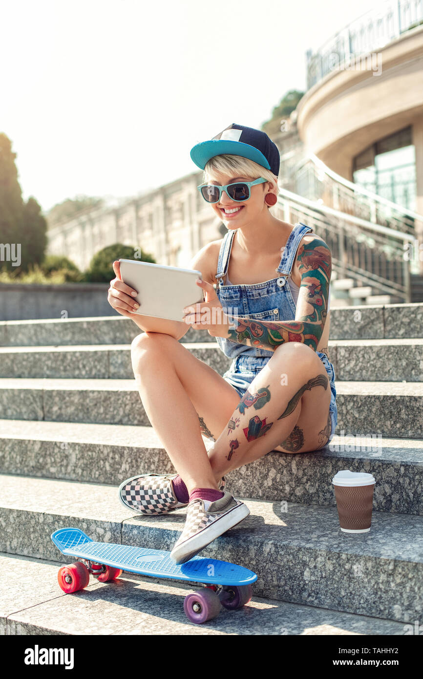 Young alternative girl skater wearing cap and sunglasses sitting on  concrete stairs with penny board and cup of hot coffee playing game online  on digi Stock Photo - Alamy