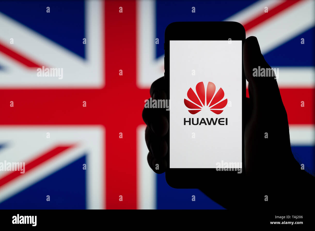 A silhouetted hand of a man holds a smartphone displaying the logo of Chinese company Huawei, with a UK flag in the background (Editorial use only). Stock Photo
