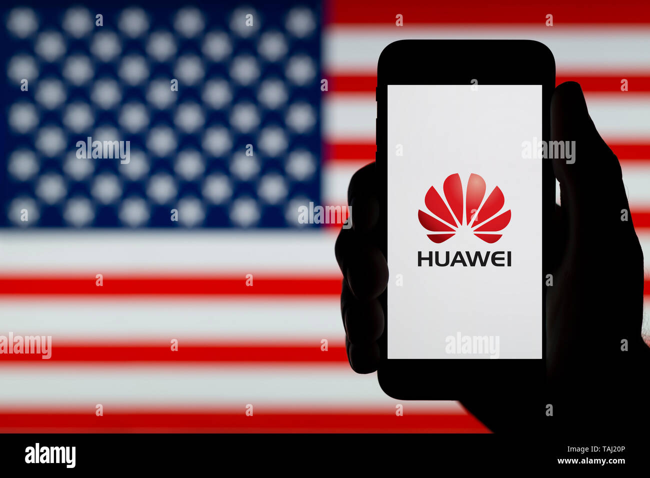 A silhouetted hand of a man holds a smartphone displaying the logo of Chinese company Huawei, with a USA flag in the background (Editorial use only). Stock Photo
