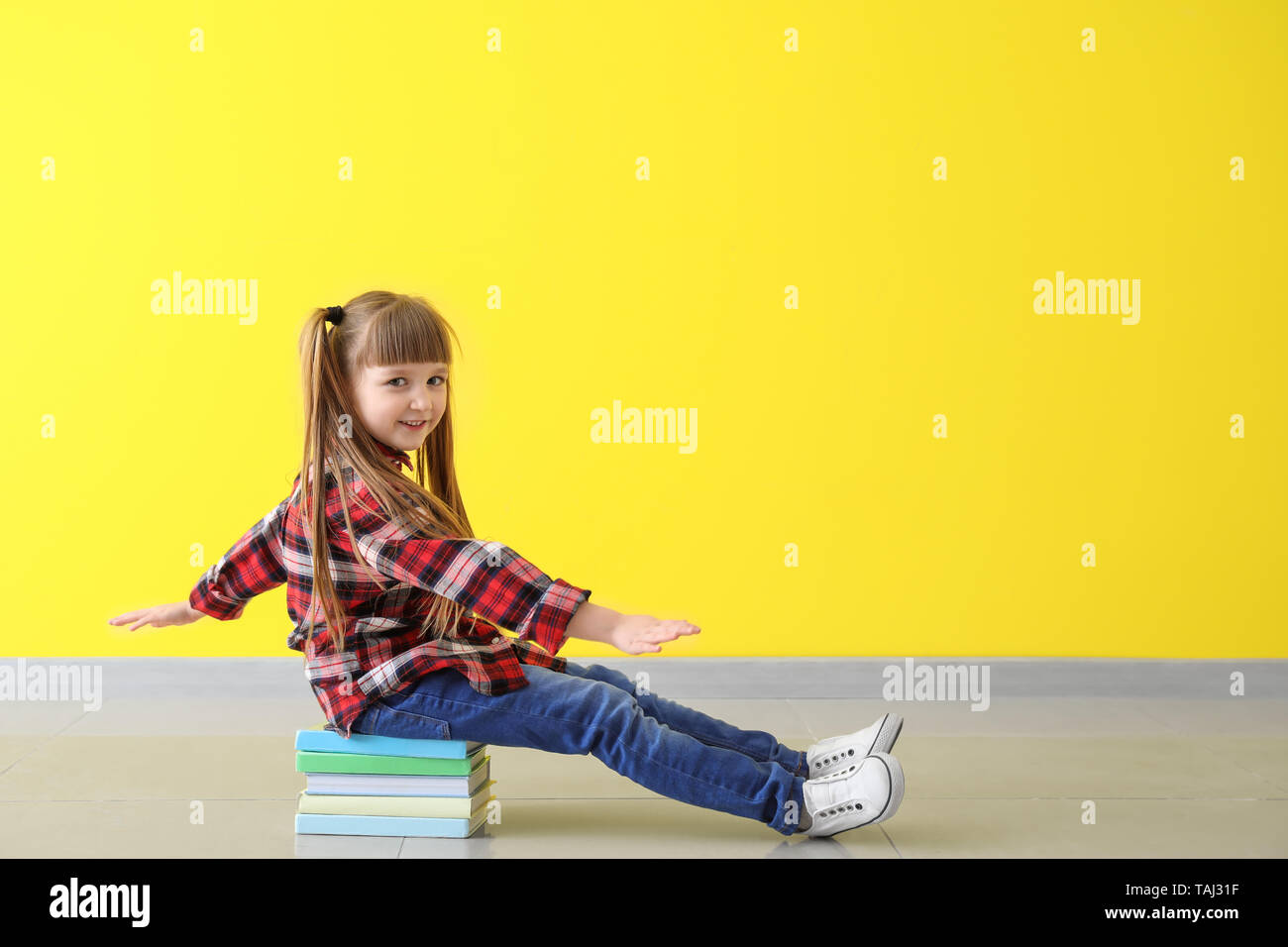 Cute little girl sitting on stack of books against color wall Stock ...