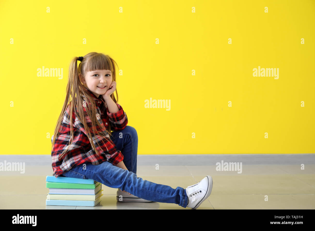 Cute little girl sitting on stack of books against color wall Stock ...