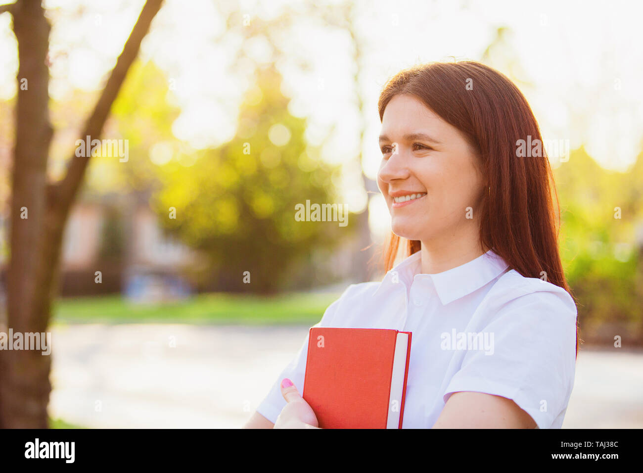 Excited smiling student girl outdoors holding a red book over sunny background . Smiling young student girl holding a book in campus. University conce Stock Photo