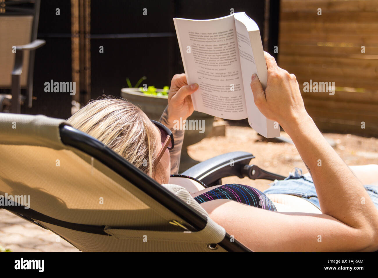blond woman reading a book and taking sunbath in the backyard at summer time Stock Photo