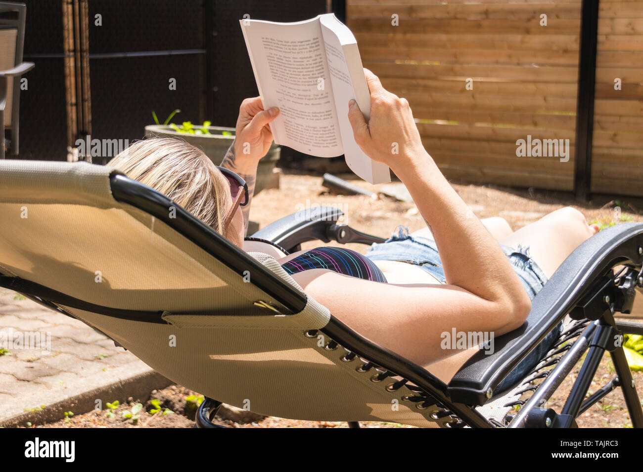blond woman reading a book and taking sunbath in the backyard at summer time Stock Photo