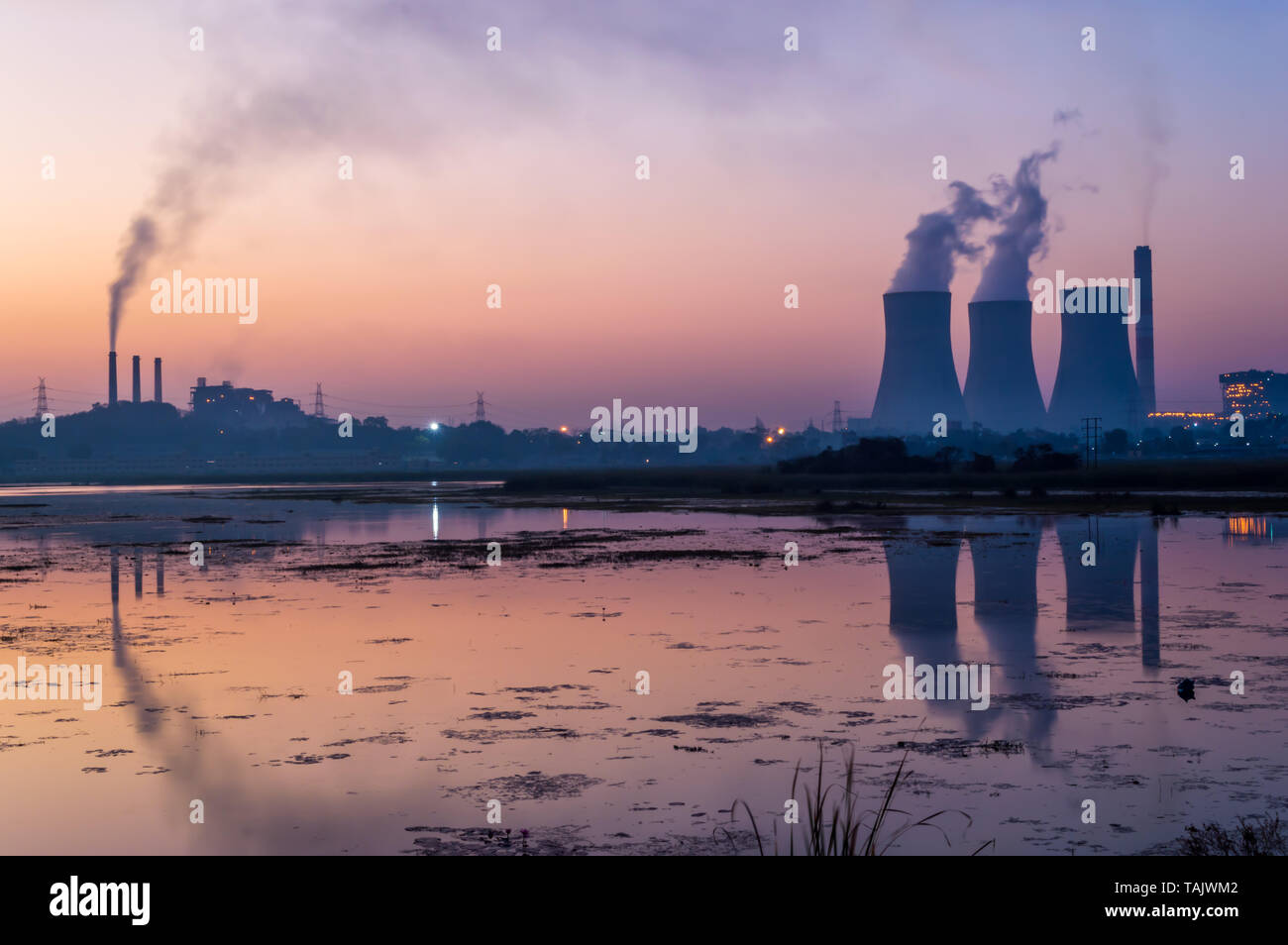 Coal powered thermal power plant emitting smoke and steam from chimney and cooling tower. Reflection of cooling tower and chimney on lake. Stock Photo