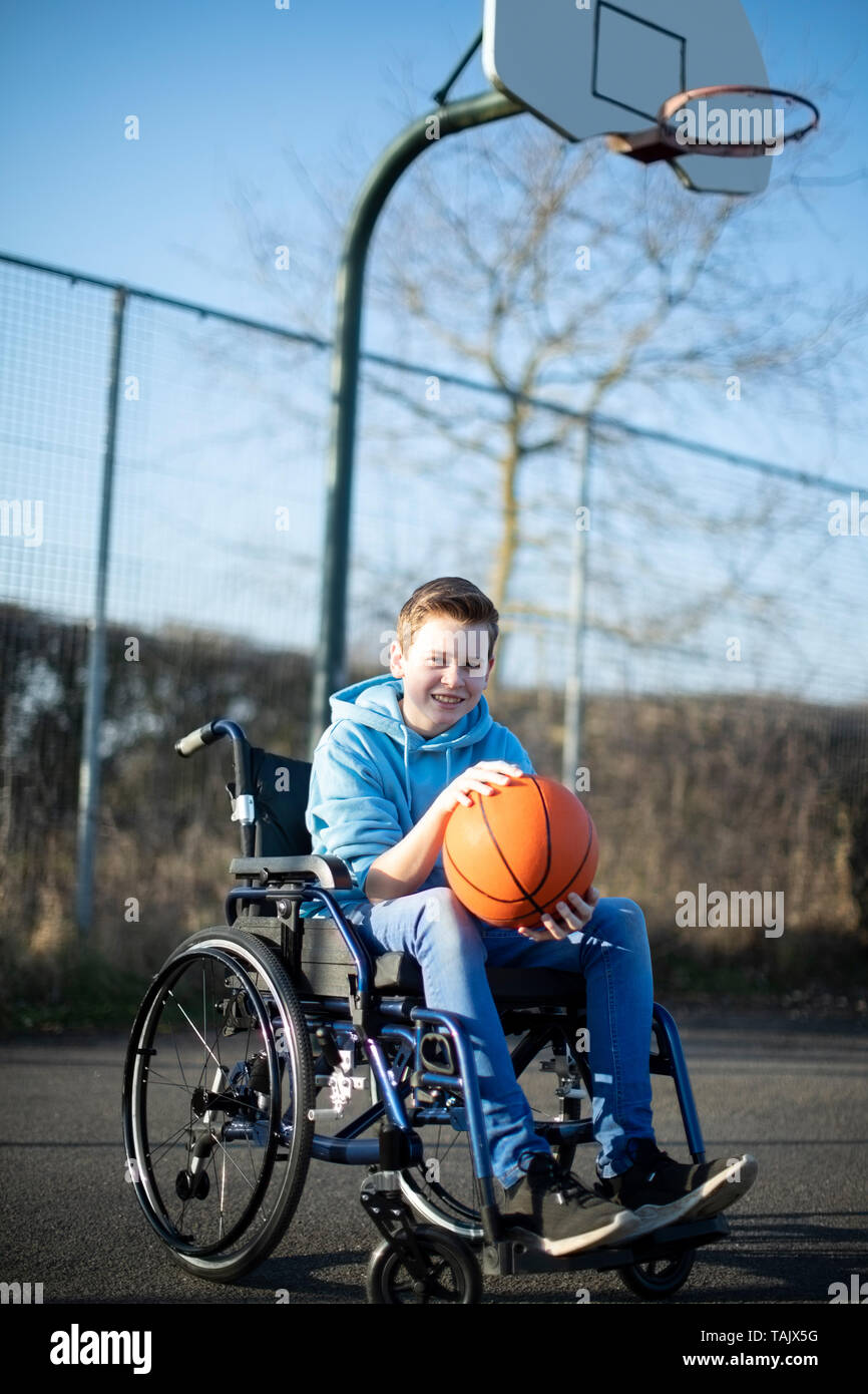 Portrait Of Teenage Boy In Wheelchair Playing Basketball On Outdoor Court Stock Photo