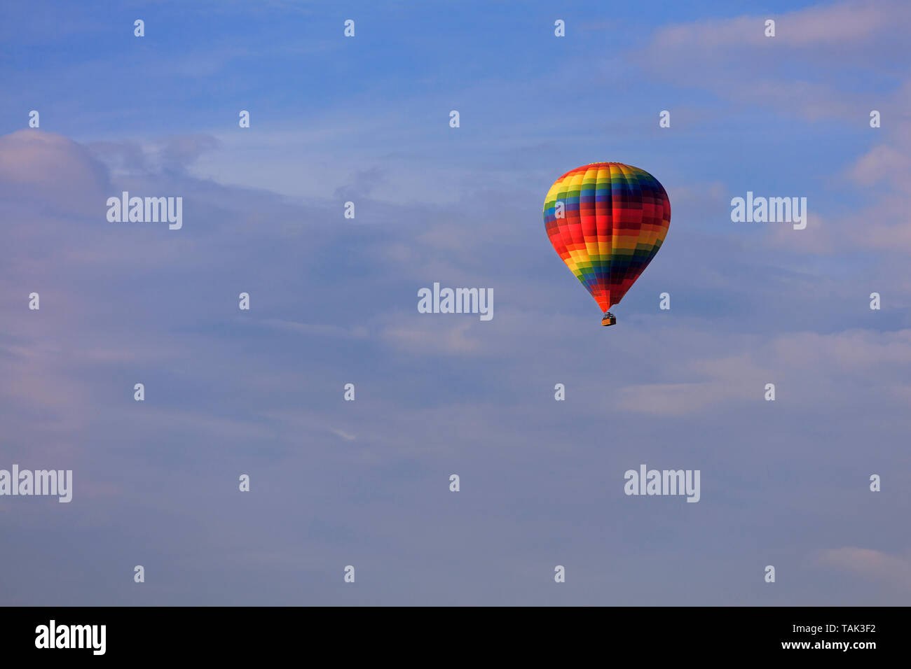 A motley multicolored beautiful balloon raises the tourist's basket to the blue sky under the sun rays among the clouds. Stock Photo