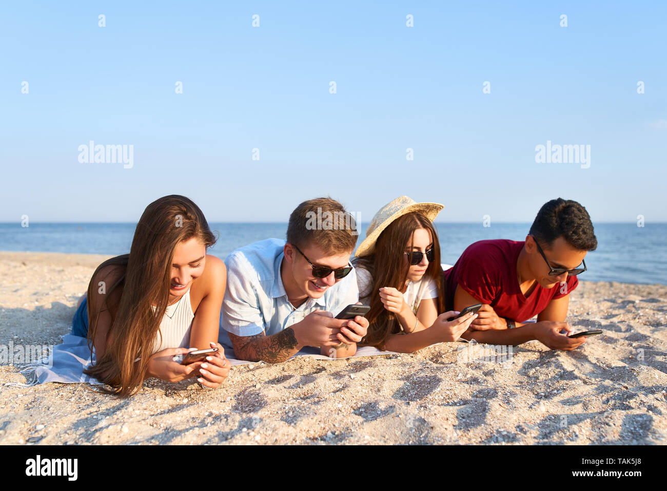 Group of millenials using smartphones laying together on beach towel near sea on summer sunset. Young people addicted by mobile smart phones. Always Stock Photo