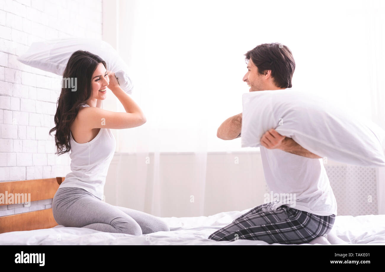 Cheerful loving couple is fighting by pillows in bed. Stock Photo
