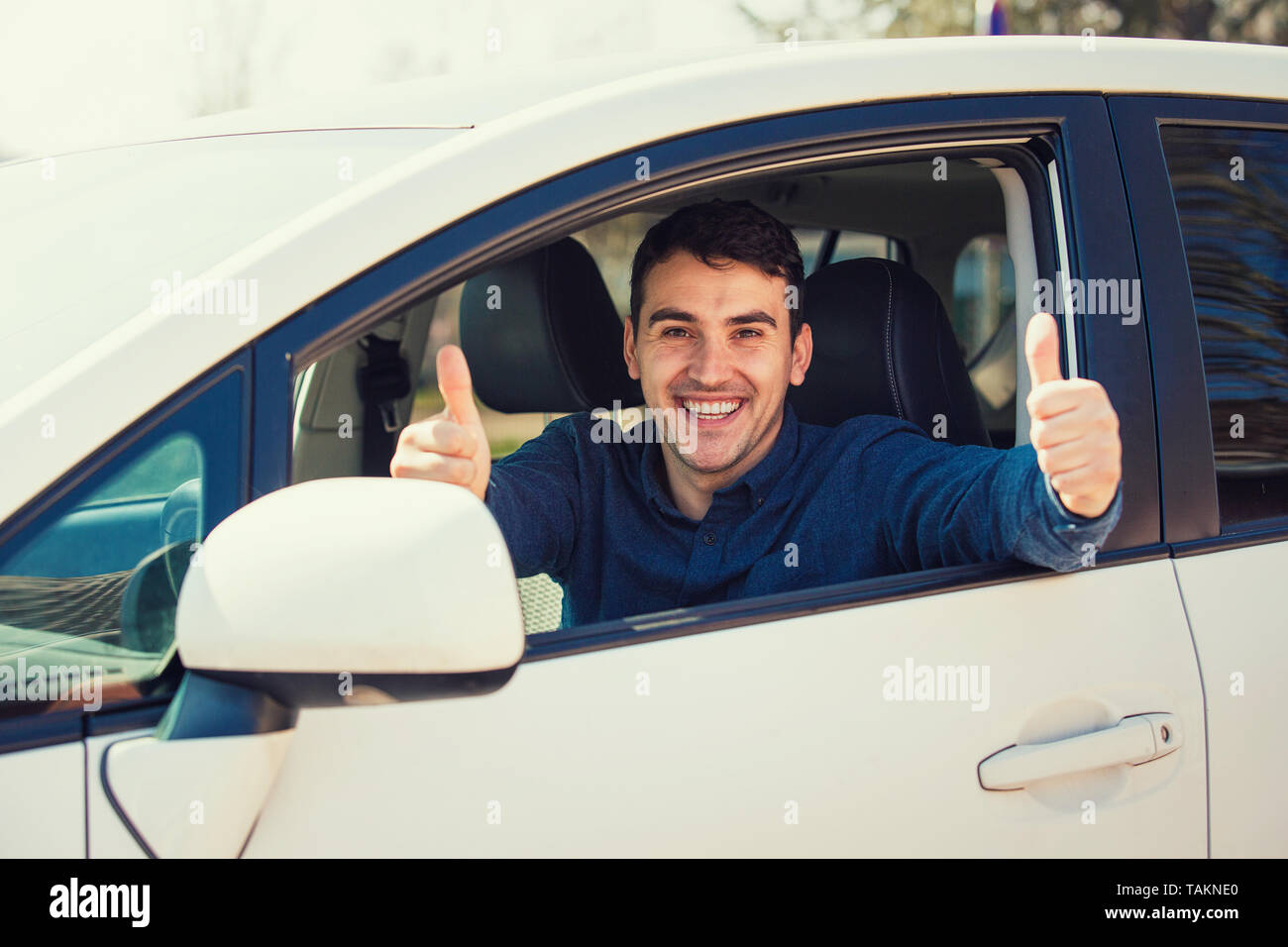Excited casual guy sitting behind the steering wheel of the car showing thumbs up gesture as has passed the exam and obtain driver license. Proud succ Stock Photo