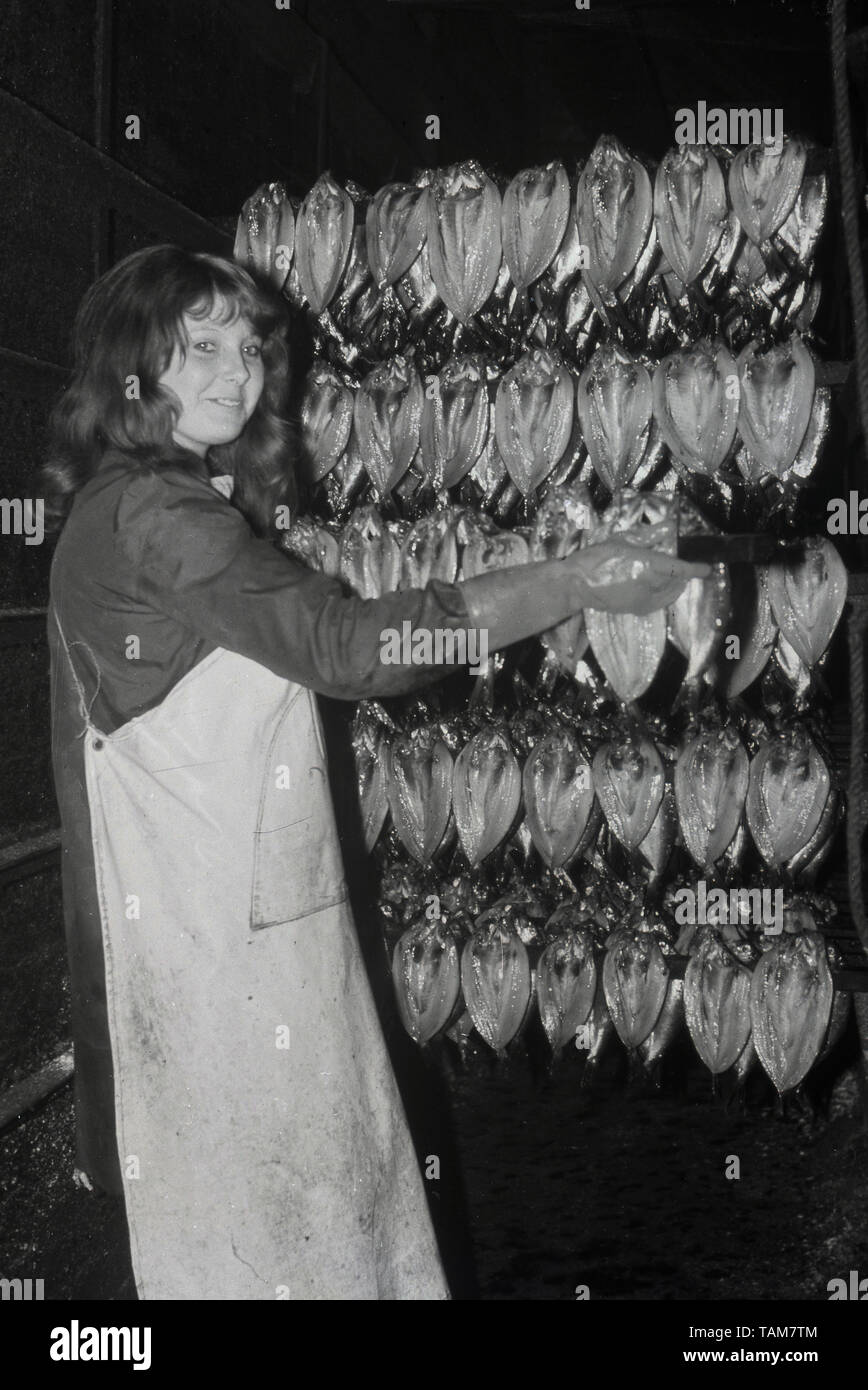 1960s, historical, young adult female worker in overalls and rubber gloves with racks of smoked kippers, England. A kipper is a whole herring, a small oily fish that has been split and cold-smoked over smouldering woodchips. Full of healthy Omega 3 oils, they are often eaten for breakfast. Stock Photo