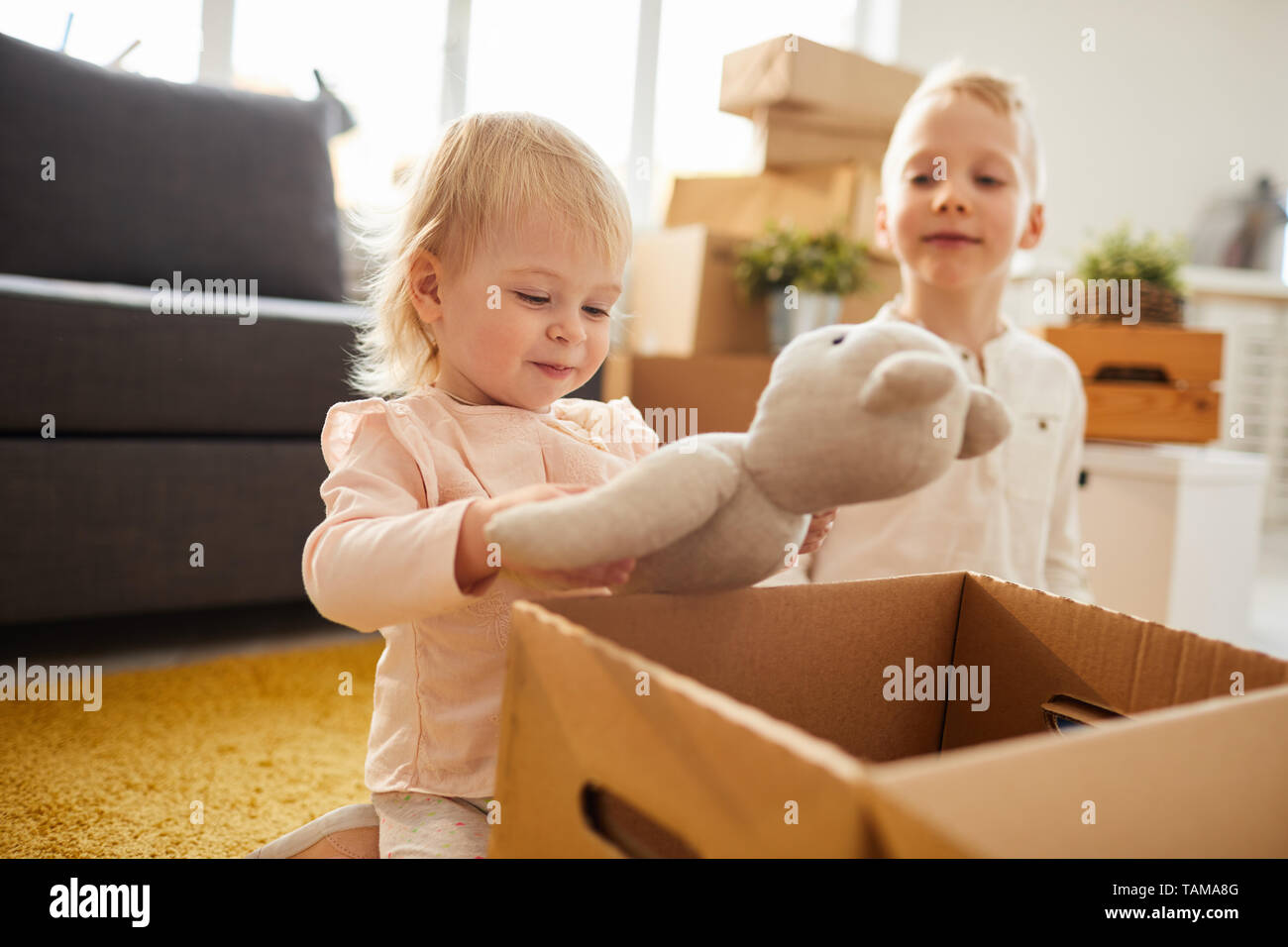 Happy cute little girl with blond hair sitting on carpet in living room of new flat and has found favorite toy in moving box, her brother being proud  Stock Photo