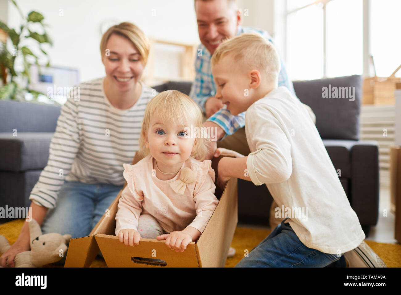 Positive playful young family with two children having fun in new flat: pretty little girl sitting in moving box and looking at camera while other fam Stock Photo