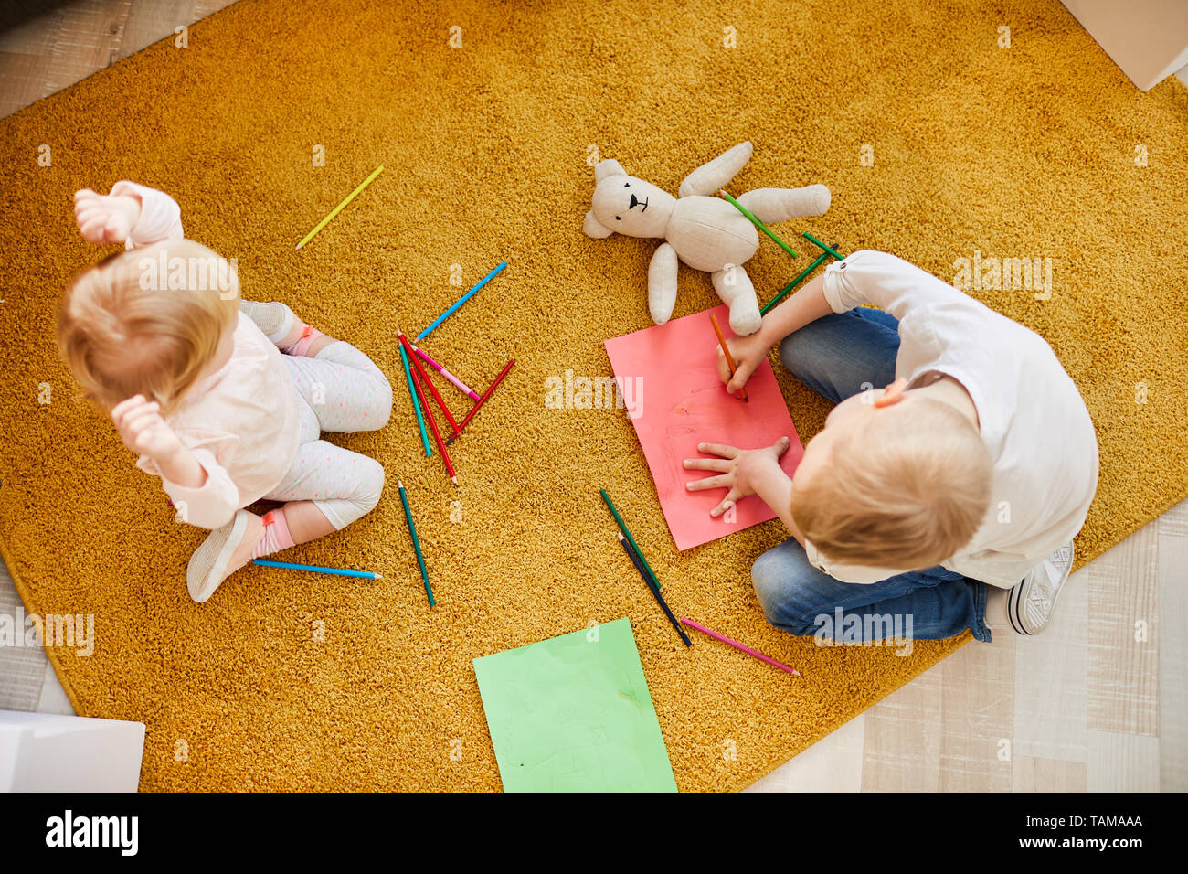 High angle view of curious kids of different ages sitting on comfortable carpet and drawing with colorful pencils on paper Stock Photo