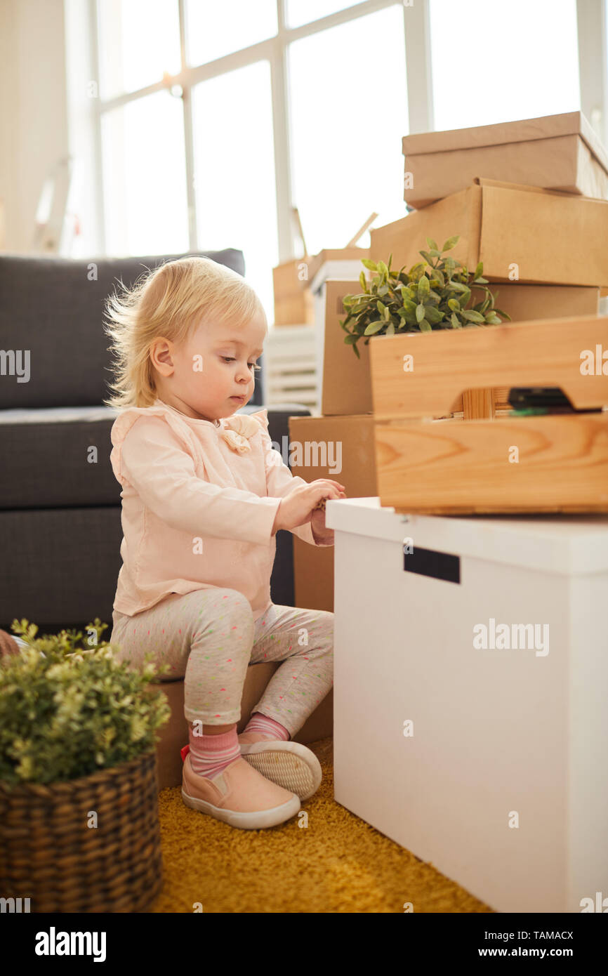 Serious curious blond-haired girl in light pink sweater sitting on box and opening moving box under stack of stuff Stock Photo