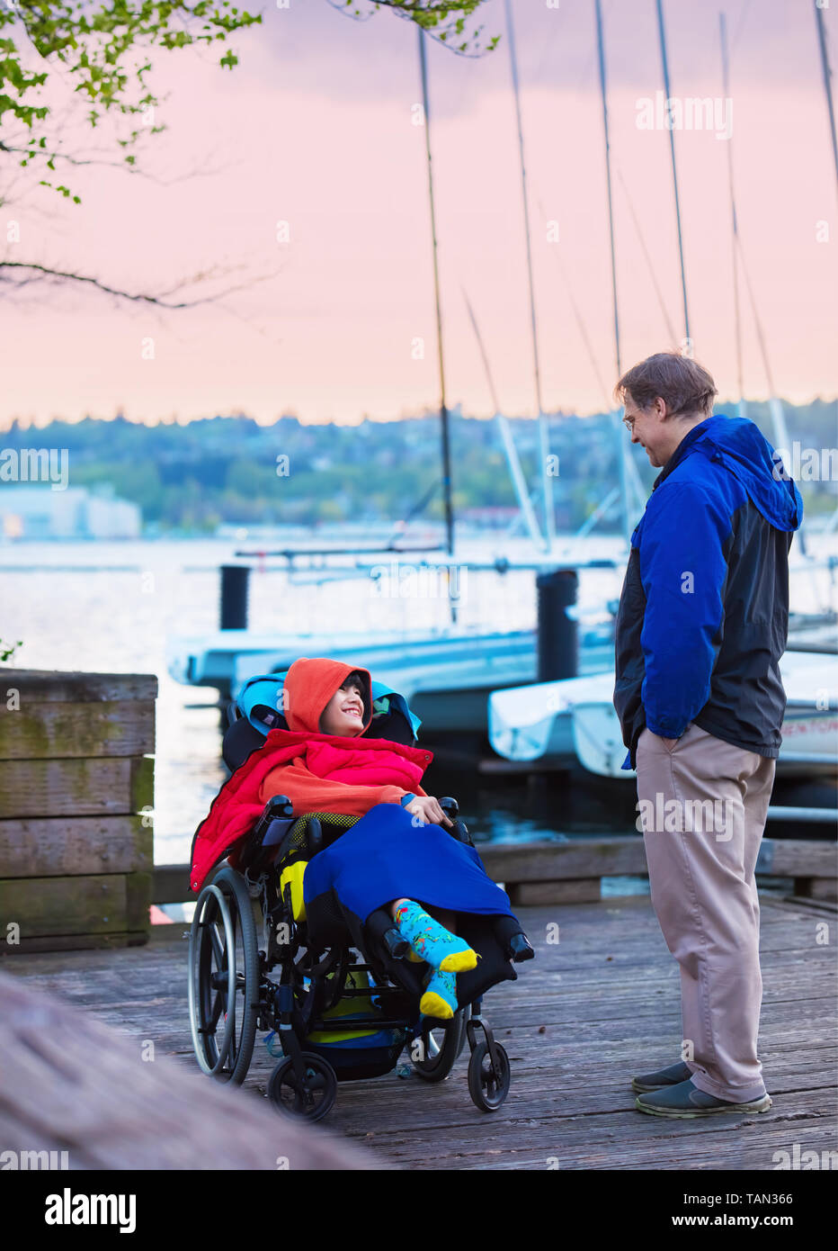 Caucasian father talking to disabled special needs son in wheelchair while standing on wooden pier near lake at sunset or dusk, small sailing boats in Stock Photo