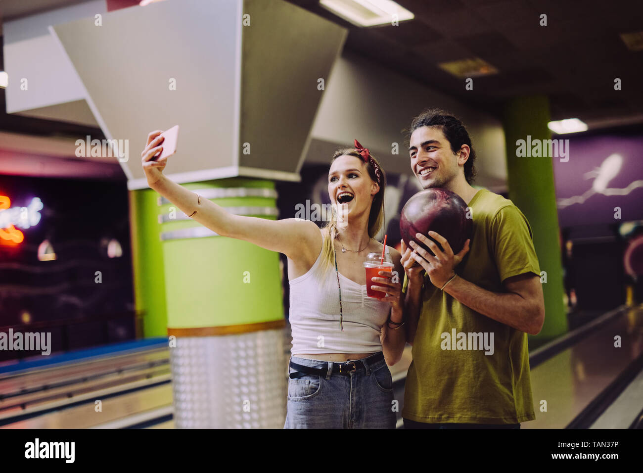 Couple taking selfie at bowling alley. Woman holding a softdrink taking selfie with man holding a ball. Stock Photo