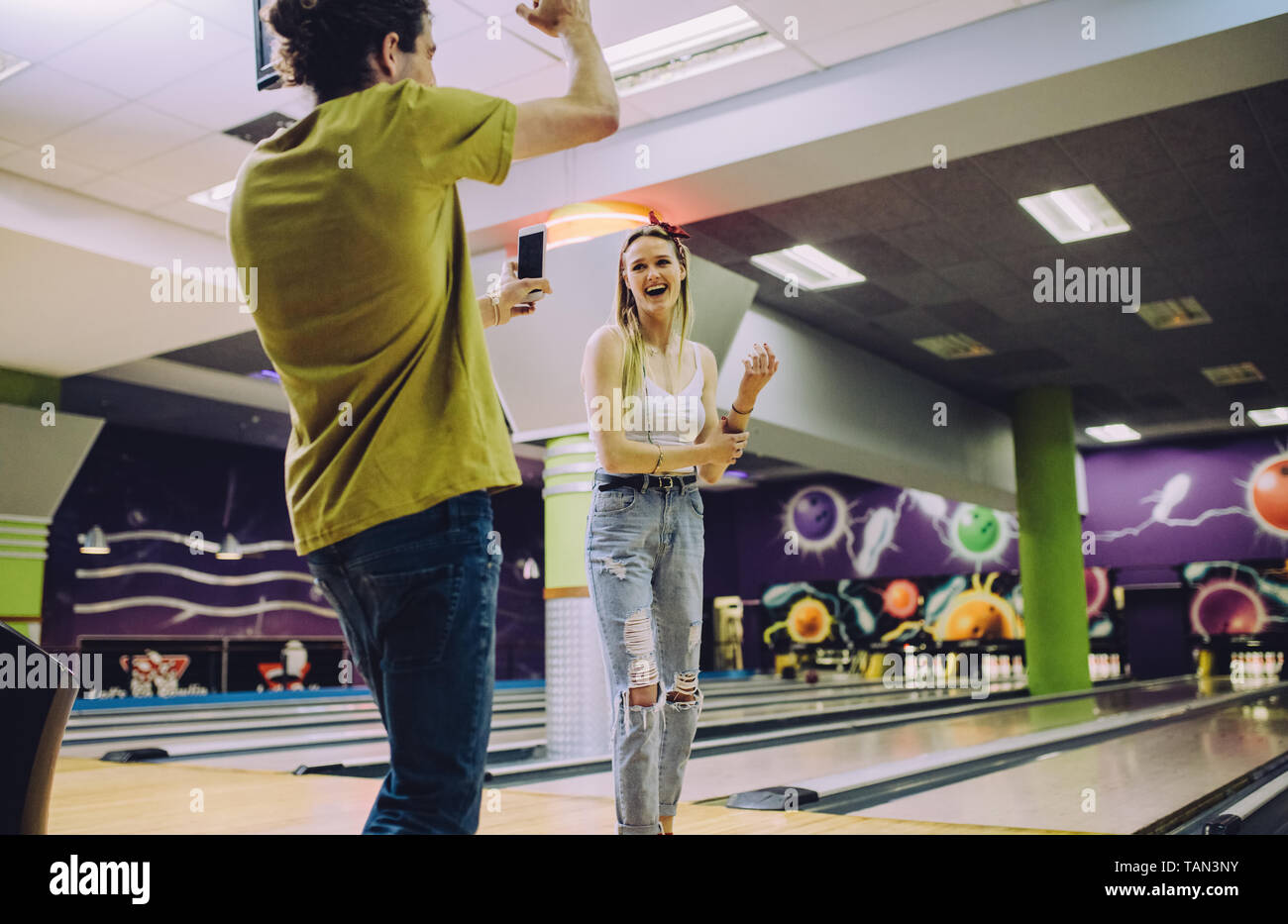 Young man recording woman playing bowling at club. Young friends enjoying playing at bowling arena. Stock Photo