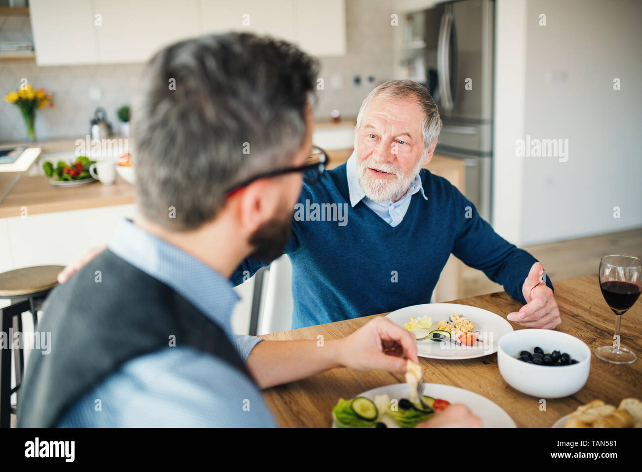 An adult hipster son and senior father indoors at home, eating light lunch. Stock Photo