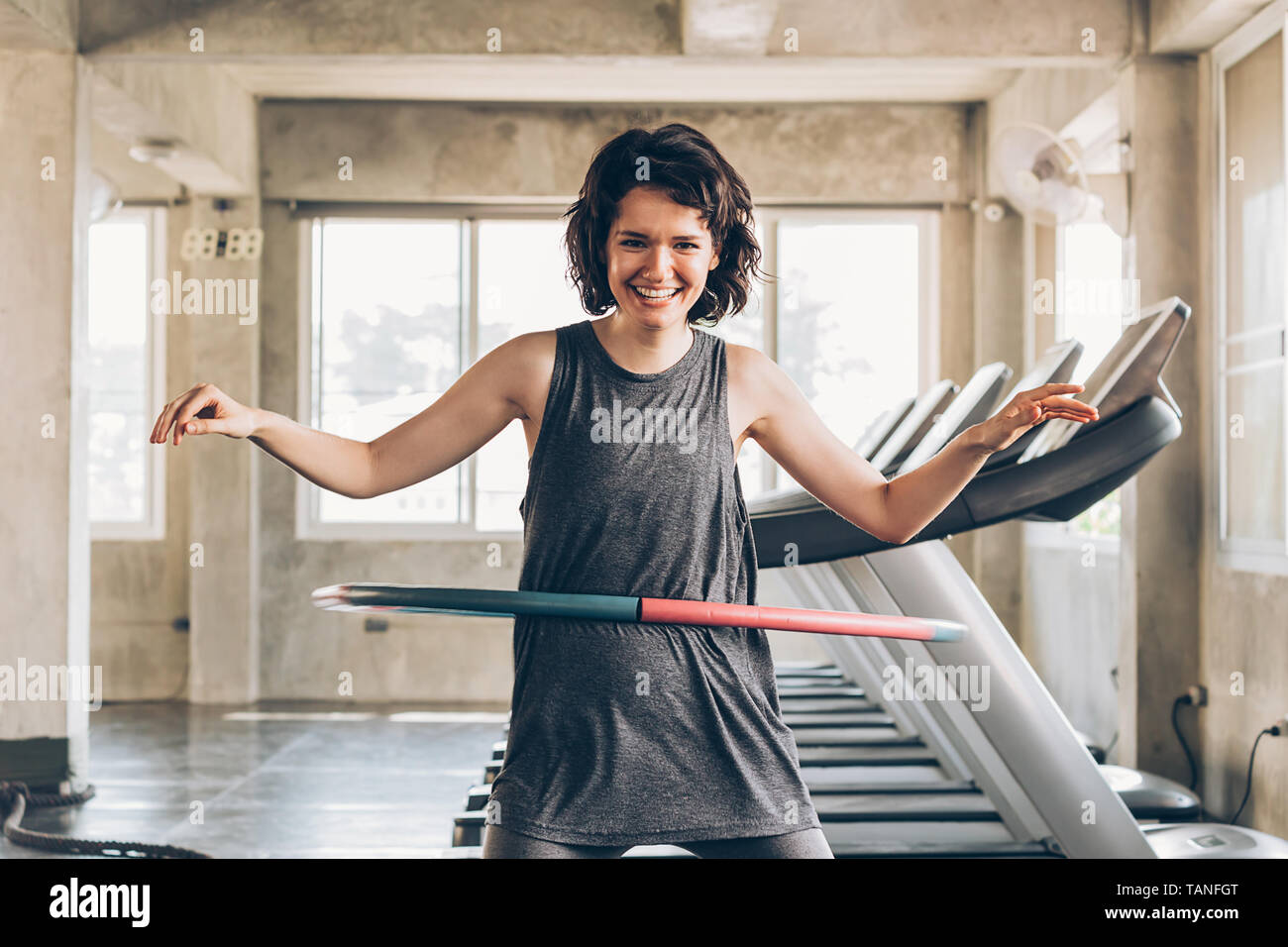 Beautiful young smiling happy Caucasian sporty woman with short hair playing hula hoop inside gym studio with treadmills behind - fun workout fitness Stock Photo
