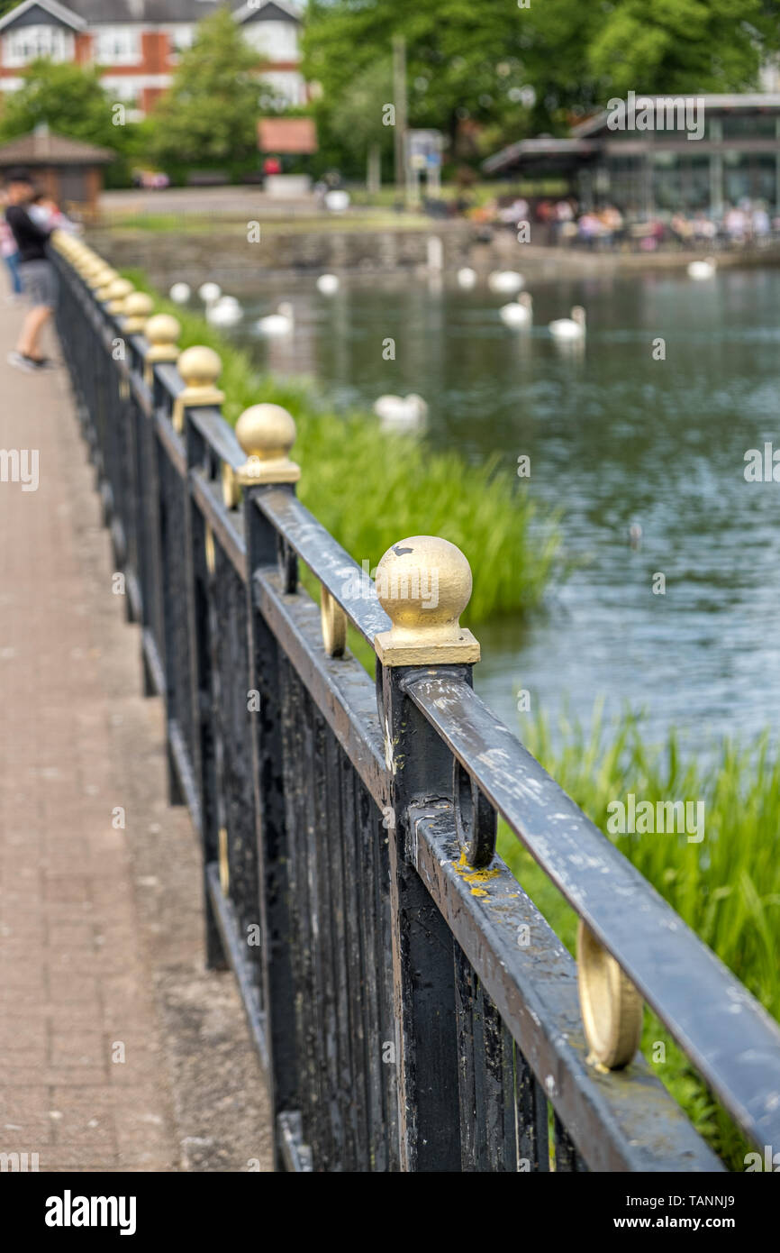 View of black iron railings topped with golden balls leading away into the distance, with a shallow depth of field. Stone walkway one side and a lake  Stock Photo