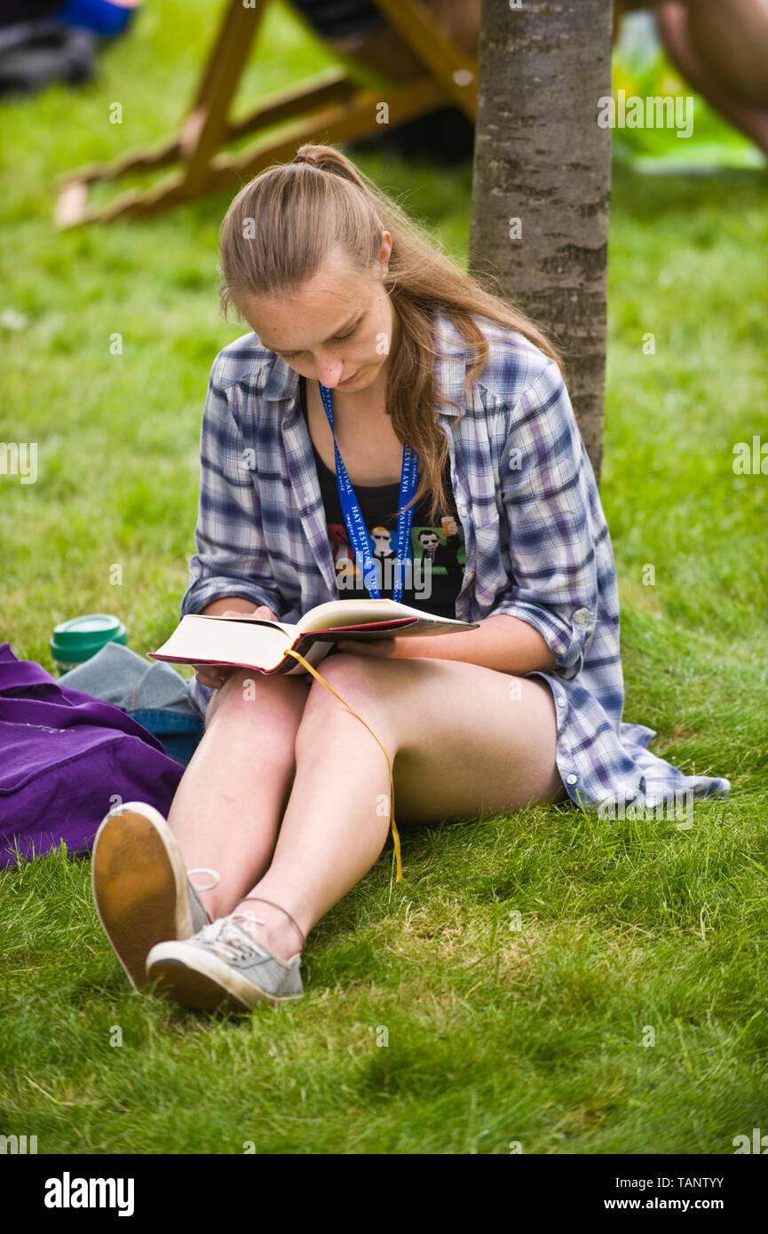 Young woman sat on grass leaning against a tree reading a book at Hay Festival Hay-on-Wye Powys Wales UK Stock Photo