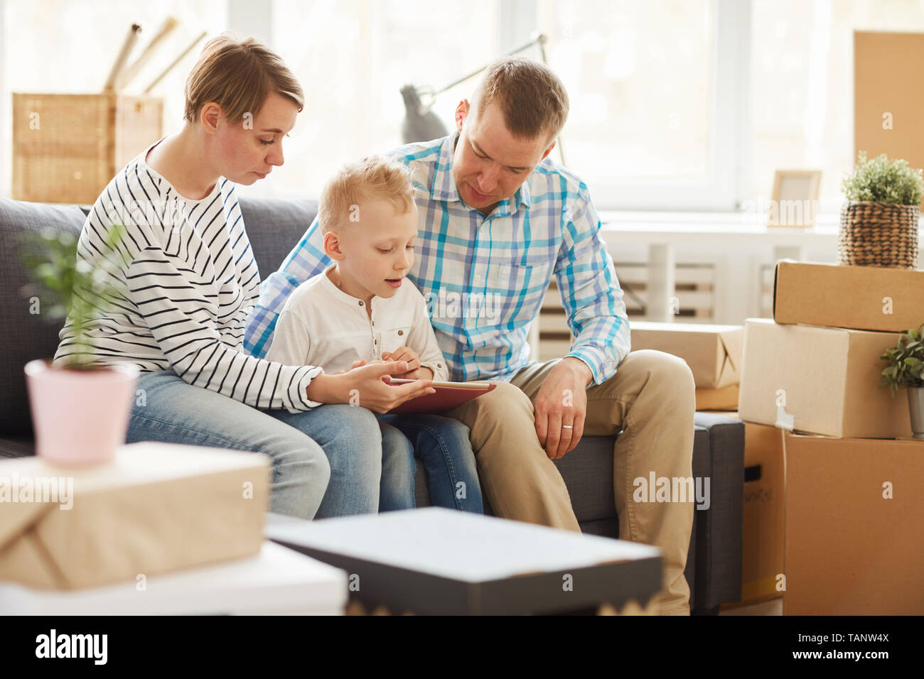 Content curious blond-haired son sitting with parents on sofa in living room with stacks of boxes and reading book on tablet while doing home task. Stock Photo