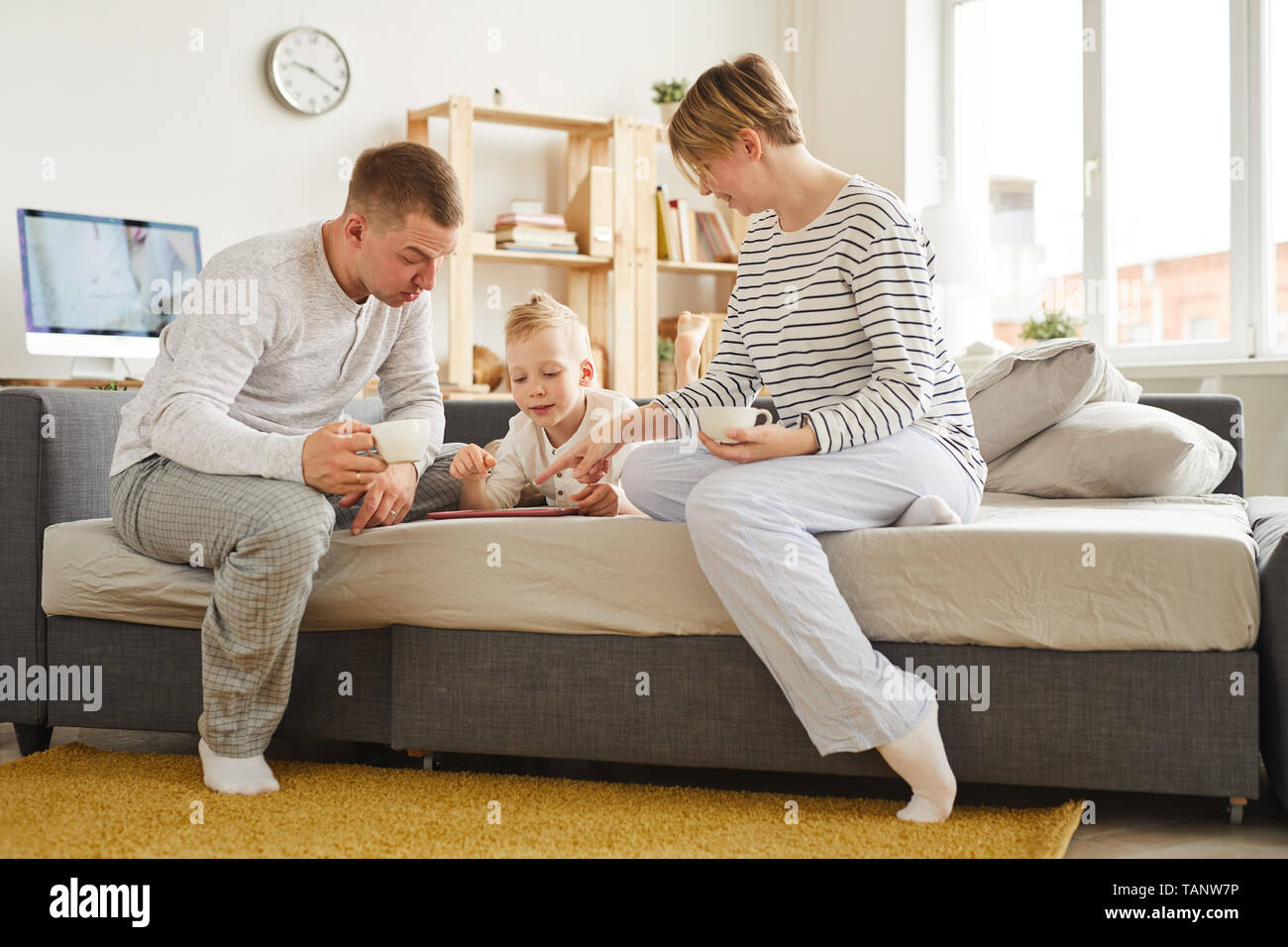 Content curious little boy lying on sofa bed and using learning app on tablet with parents, young mother and father drinking coffee and helping son Stock Photo