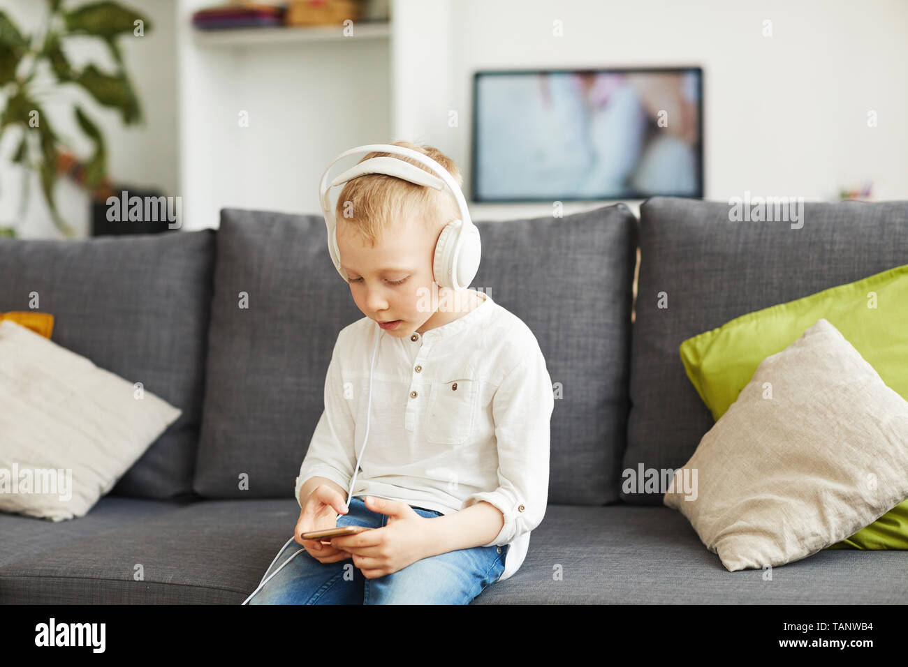 Content curious technology-addicted little boy in headphones sitting on sofa and playing game on phone Stock Photo