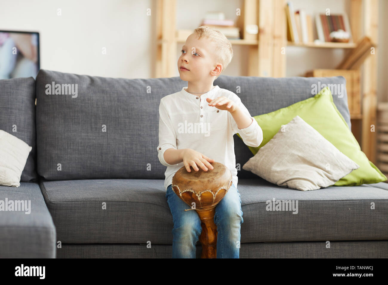 Pensive creative Caucasian boy with blond hair sitting on comfortable sofa in living room and hitting drum with hands while playing djembe Stock Photo