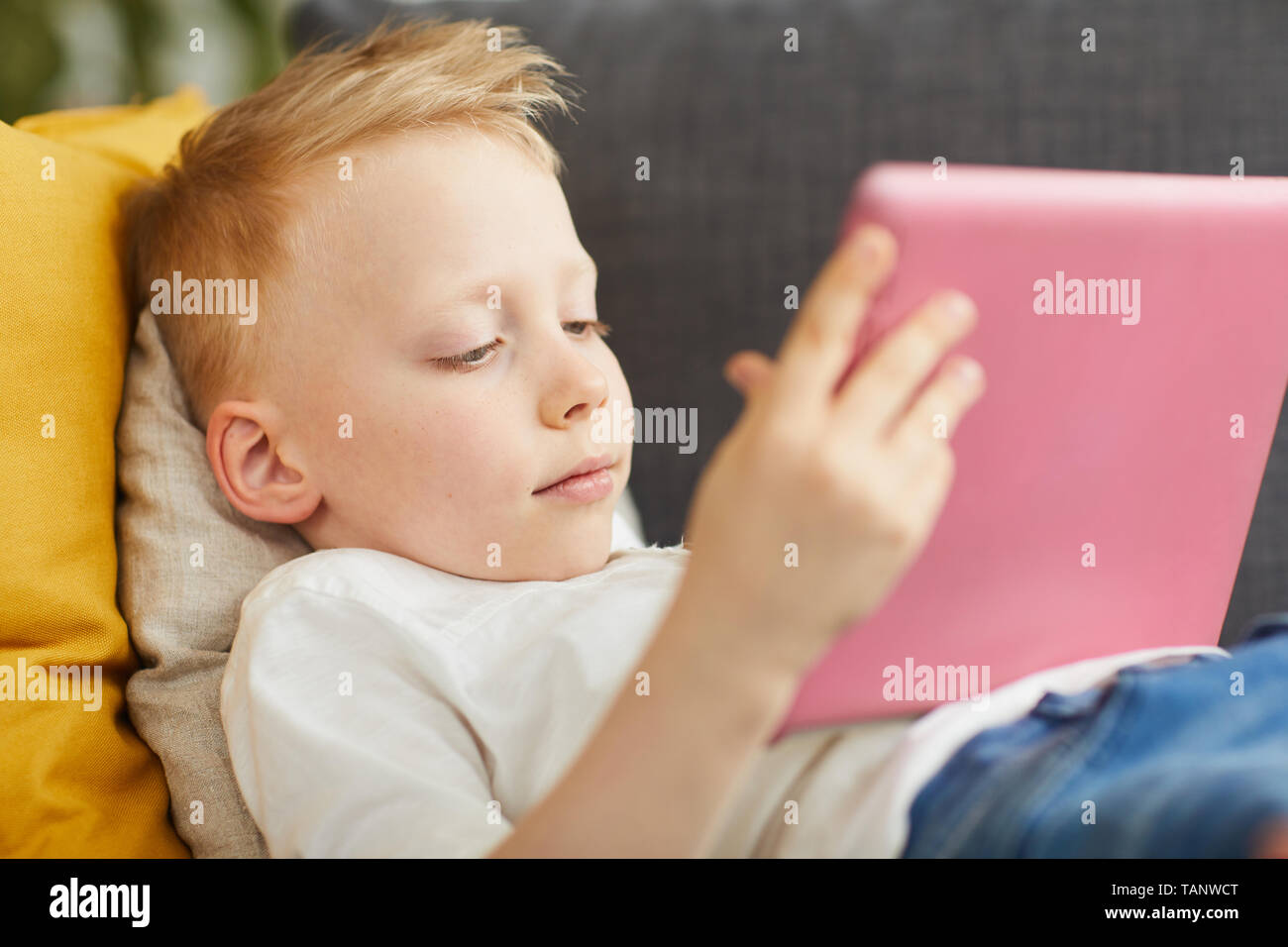 Close-up of serious curious little boy with blond hair lying on pillows and using tablet while watching educational video at home Stock Photo