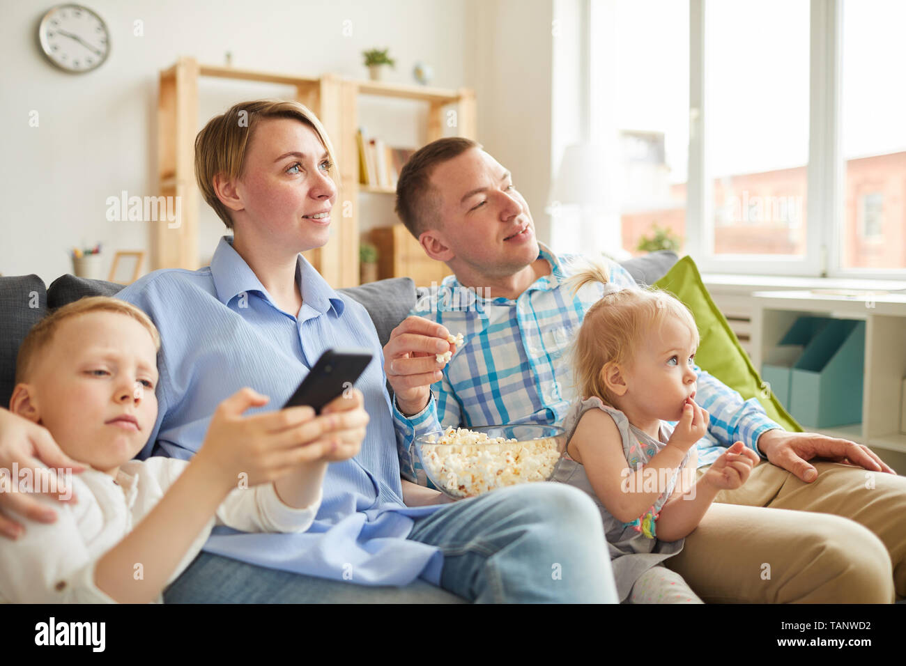 Concentrated curious young family sitting on sofa in living room and watching tv while eating popcorn, they spending leisure in front of television se Stock Photo