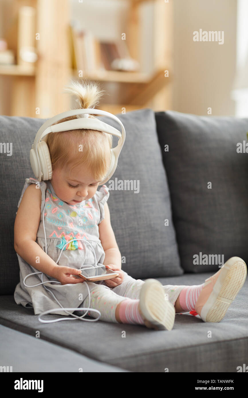 Curious cute toddler girl in gray dress sitting on comfortable sofa and using mobile app on smartphone while listening to music in wired headphones Stock Photo