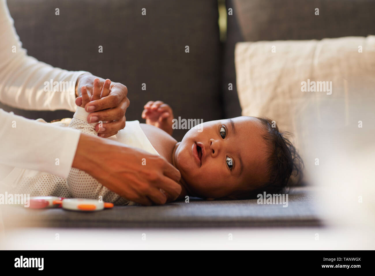 Cropped image of cute black baby boy with chubby cheeks lying on sofa and looking at camera while mother dressing him Stock Photo