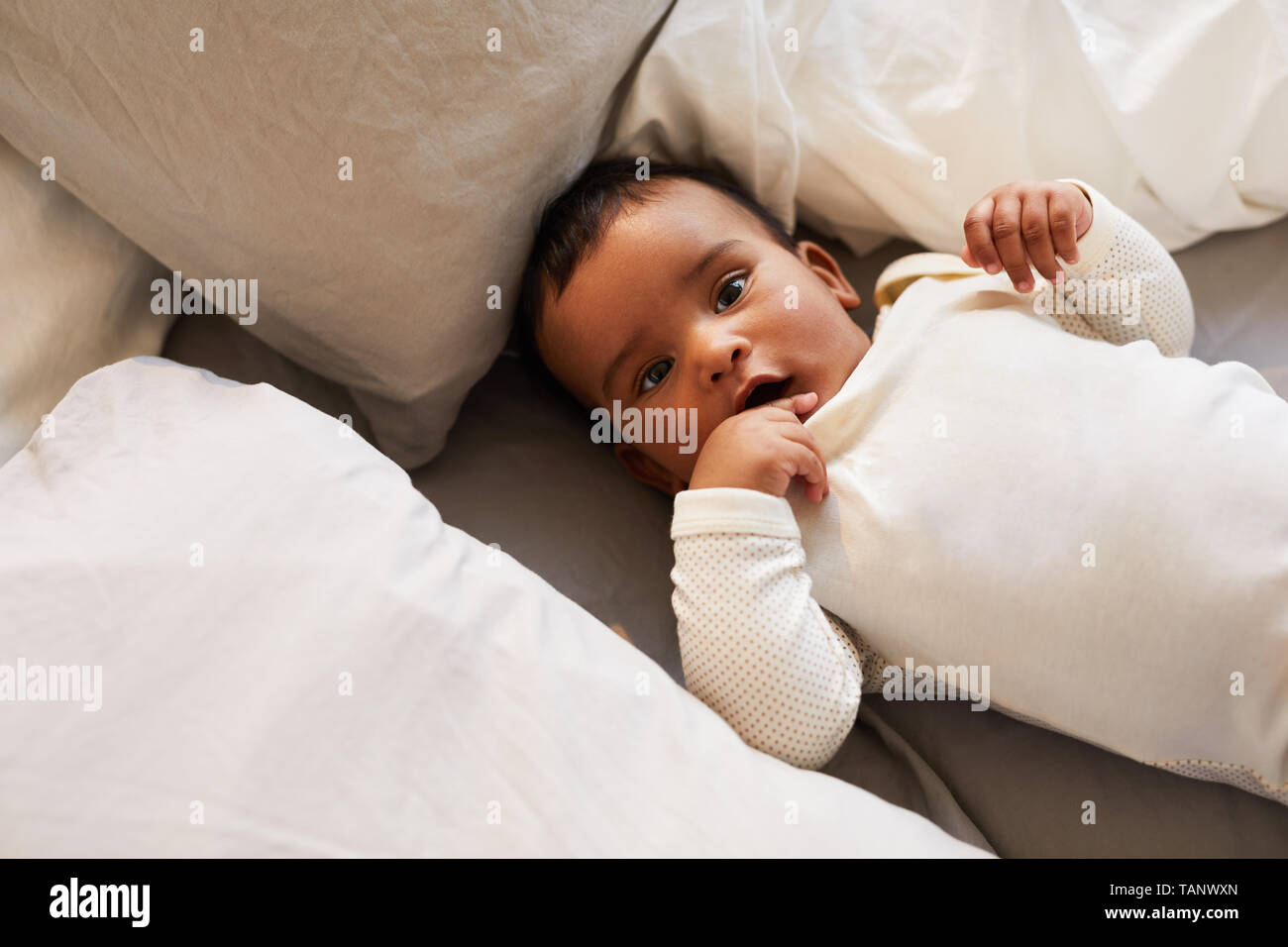 Directly above view of content curious cute baby boy in white baby suit lying in bed among soft pillows and looking at camera Stock Photo